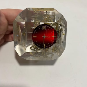 May include: A clear, cube-shaped paperweight with a red clock face and gold gears inside. The clock face has the text "24 Hours" printed on it.