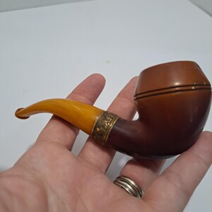 Op de afbeelding: Een bruine en amberkleurige pijp met een gouden band. De pijp wordt in een hand gehouden.