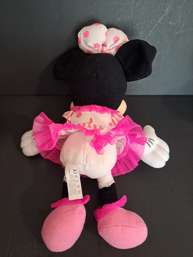 May include: Pink and black plush Minnie Mouse toy wearing a pink polka dot dress and a white bow with pink polka dots. The toy has a pink tulle skirt and pink shoes.