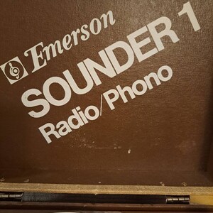 Pu&ograve; includere: Un giradischi vintage Emerson Sounder 1 Radio/Phono di colore marrone con un logo e testo bianchi.