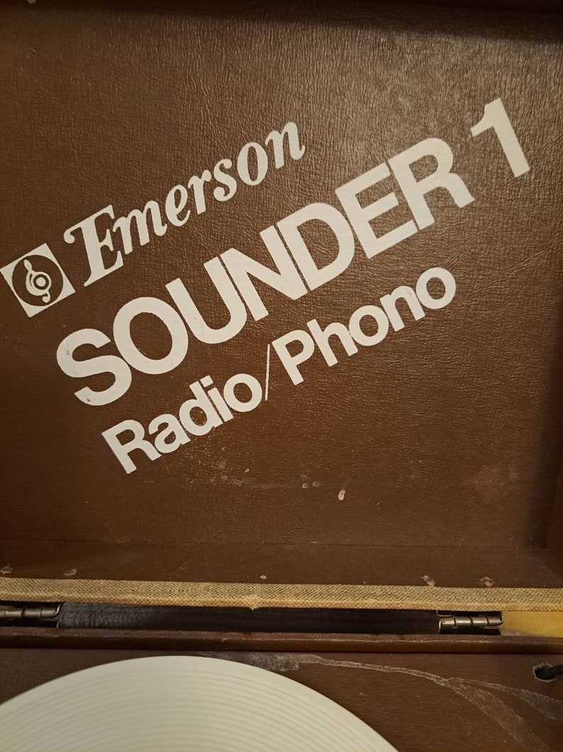 Pu&ograve; includere: Un giradischi vintage Emerson Sounder 1 Radio/Phono di colore marrone con un logo e testo bianchi.