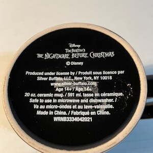 May include: A black ceramic mug base featuring white text that reads Disney Tim Burton's The Nightmare Before Christmas, Silver Buffalo, LLC, New York, NY 10018, 20 oz. ceramic mug, safe to use in microwave and dishwasher, Made in China, WRNB3334042021.