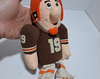May include: A brown and orange plush football player doll wearing a helmet and jersey with the number 19. The doll has a large nose and a friendly smile.
