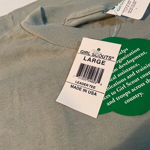 May include: A sage green t-shirt with a crew neckline and a white rectangular tag attached. The tag reads GIRL SCOUTS LARGE, LEADER TEE, MADE IN USA, and includes a barcode. A green clover-shaped tag is also attached with printed text.