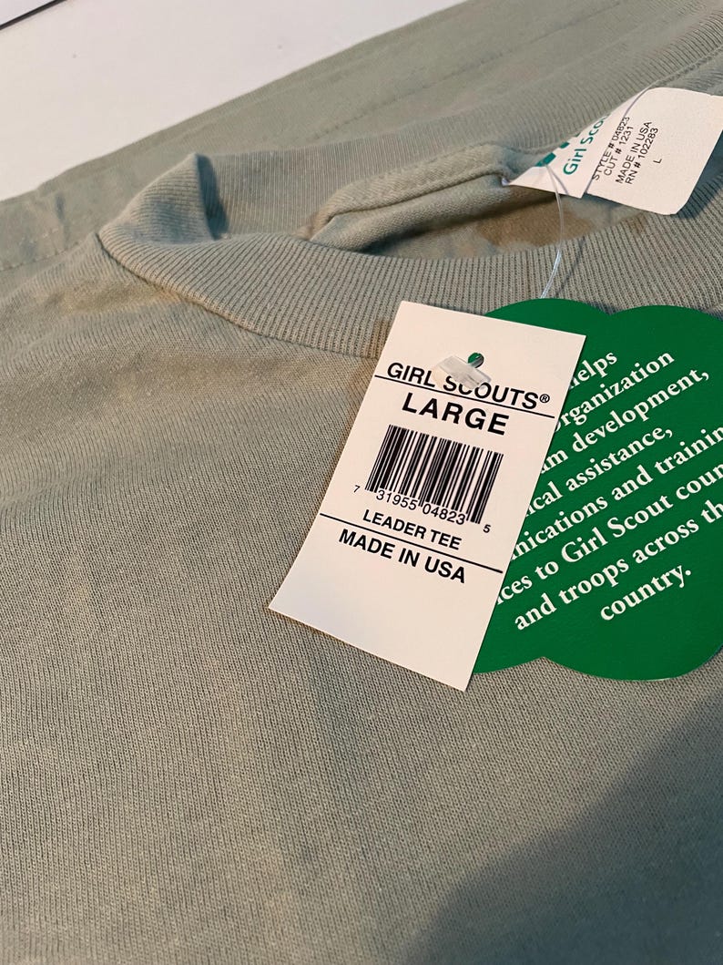 May include: A sage green t-shirt with a crew neckline and a white rectangular tag attached. The tag reads GIRL SCOUTS LARGE, LEADER TEE, MADE IN USA, and includes a barcode. A green clover-shaped tag is also attached with printed text.