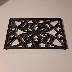 May include: A black cast iron trivet with a decorative, intricate design. The trivet is rectangular and has a raised edge.
