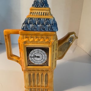 May include: A ceramic teapot shaped like the Big Ben clock tower. The teapot is primarily yellow with blue and gold accents. The clock face is visible on the front. The handle and spout are also yellow.