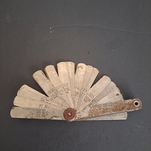 May include: A metal feeler gauge with a fan-shaped design. The gauge has a series of blades with different thicknesses, marked with numbers from 0.035 to 0.220 inches.