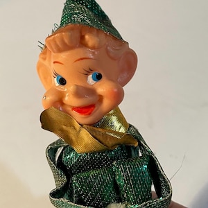 May include: Vintage Christmas elf ornament with a smiling face, blue eyes, and a red mouth. The elf wears a green metallic outfit with a matching hat and gold accents. A festive holiday decoration.