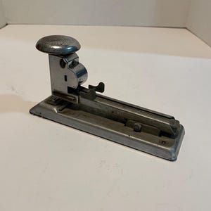 May include: A vintage silver metal stapler with a large round top and a long, flat base. The stapler has a lever on the side and a small metal plate on the front.