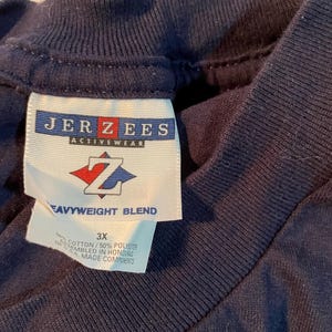 K&ouml;nnte beinhalten: Ein dunkelblaues T-Shirt mit einem wei&szlig;en Innenetikett am Kragen, das das Jerzees Activewear-Logo, ein rot-blaues Z-Emblem und den Text Heavyweight Blend zeigt. Das Etikett gibt die Gr&ouml;&szlig;e 3X und eine Mischung aus 50% Baumwolle und 50% Polyester an.