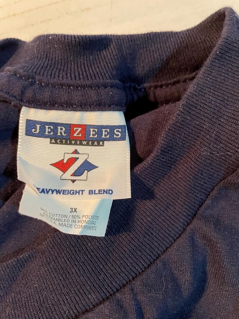 K&ouml;nnte beinhalten: Ein dunkelblaues T-Shirt mit einem wei&szlig;en Innenetikett am Kragen, das das Jerzees Activewear-Logo, ein rot-blaues Z-Emblem und den Text Heavyweight Blend zeigt. Das Etikett gibt die Gr&ouml;&szlig;e 3X und eine Mischung aus 50% Baumwolle und 50% Polyester an.