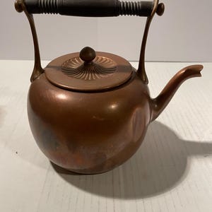 May include: A copper kettle with a rounded body, a curved spout, and a removable lid featuring a decorative fluted design. The kettle has a tall, arched metal handle topped with a dark, ribbed cylindrical grip for handling.