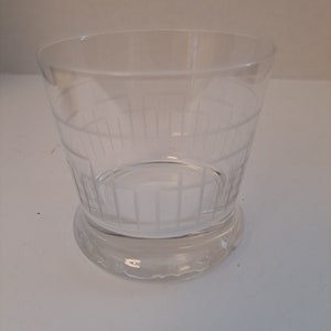 May include: A clear, etched glass tumbler with a wide, cylindrical shape and a rounded base. The glass features vertical and horizontal etched lines, creating a grid-like pattern around the body of the glass. The glass sits on a clear, circular base.