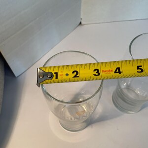 May include: Clear glass tumbler with a measurement of approximately 3 inches in diameter. The glass has a slightly flared shape and a logo at the base. A yellow measuring tape is placed across the top of the glass.
