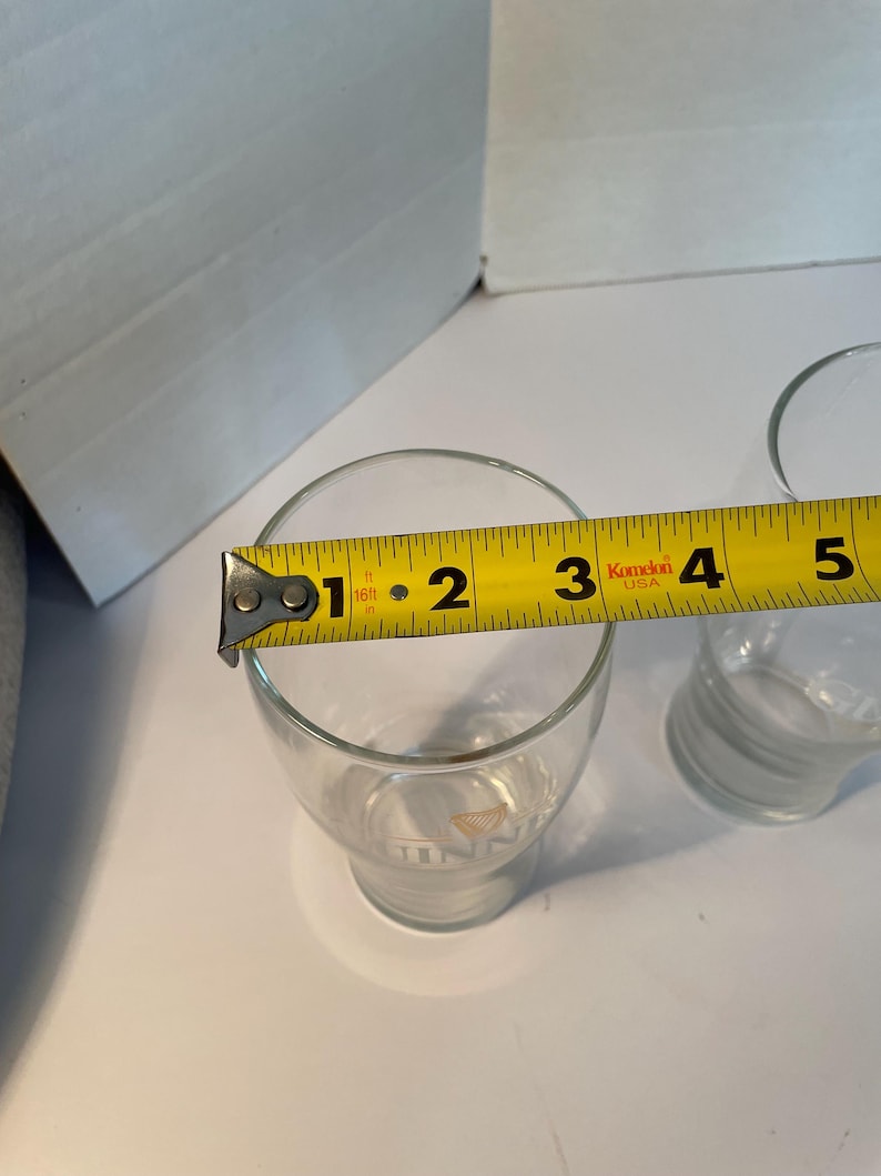 May include: Clear glass tumbler with a measurement of approximately 3 inches in diameter. The glass has a slightly flared shape and a logo at the base. A yellow measuring tape is placed across the top of the glass.
