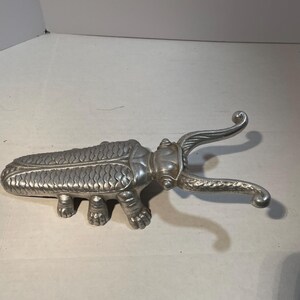 May include: A silver-colored, beetle-shaped bottle opener. The opener has a detailed design, including textured wings and legs. The beetle's pincers are curved and extend outward, designed for removing bottle caps.
