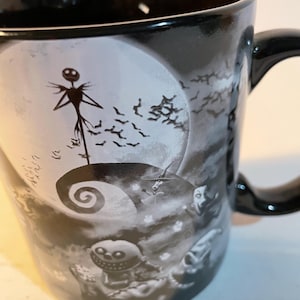 May include: A black ceramic mug featuring a monochromatic graphic print of a tall, thin skeletal figure standing on a spiral hill against a large full moon, surrounded by flying bats and other small character figures in a dark, atmospheric scene.