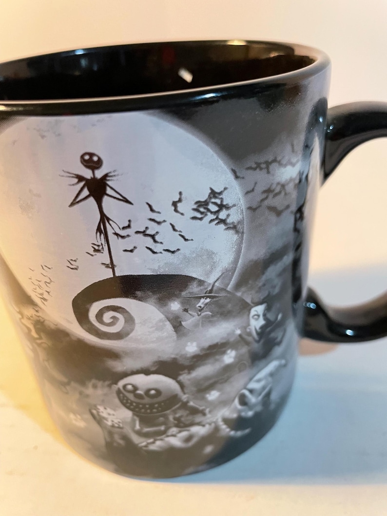May include: A black ceramic mug featuring a monochromatic graphic print of a tall, thin skeletal figure standing on a spiral hill against a large full moon, surrounded by flying bats and other small character figures in a dark, atmospheric scene.