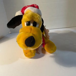 May include: A plush yellow dog toy with black ears and a black nose, wearing a red and white knit hat and a red scarf. The dog has black eyes and is sitting down. A tag is attached to the toy.