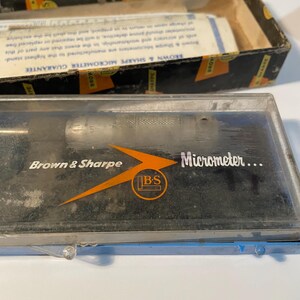 May include: A vintage Brown & Sharpe micrometer in a clear plastic case, with the brand name and "Micrometer" printed on the case. The case sits in front of a cardboard box with the brand name and guarantee information.