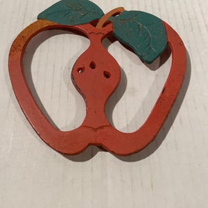 May include: A red, apple-shaped metal trivet with a cutout center design. The trivet features two dark teal leaves at the top and shows signs of wear with visible rust-colored spots and distressed paint throughout the red and green surfaces.