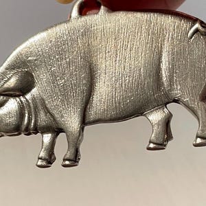May include: A silver-toned metal pendant shaped like a pig in profile. The surface features a brushed, textured finish with visible vertical lines. A small circular loop is attached to the top of the pig's back for hanging, and a small curly tail is visible.