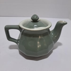 May include: A green ceramic teapot with a white lid and a white rim. The teapot has a rounded body and a spout on the side.