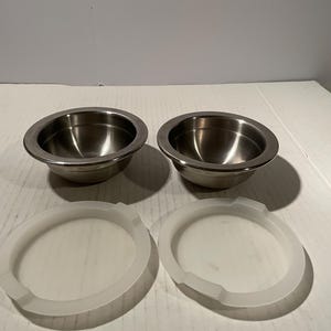 May include: Two circular stainless steel bowls with a polished metallic finish and two separate translucent white plastic lids. The bowls feature a wide rim and a deep basin, while the lids are designed to fit the circumference of the bowls.