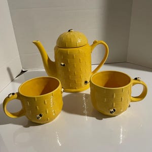 May include: A yellow ceramic teapot and two matching mugs. The teapot has a beehive design with a lid and a handle. The mugs and teapot are decorated with small bee figurines. The set is designed for serving tea.