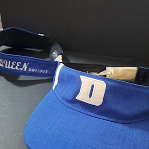 May include: Blue visor with a white "D" embroidered on the front. The visor has a white band with the text "QUEEN DRI-FIT" on it.