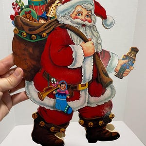 May include: A vintage wooden Santa Claus cutout with a red suit, white beard, and a bag full of toys. He is holding a toy soldier in his right hand and a candy cane in his left.