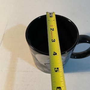 May include: A black ceramic mug with a handle, positioned beneath a yellow Stanley measuring tape that indicates a diameter of approximately 3.5 inches across the top opening of the mug. The mug features a partial graphic design on its exterior.