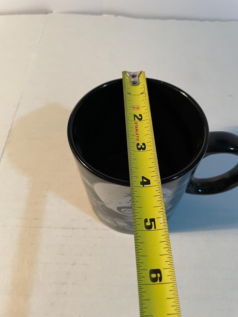 May include: A black ceramic mug with a handle, positioned beneath a yellow Stanley measuring tape that indicates a diameter of approximately 3.5 inches across the top opening of the mug. The mug features a partial graphic design on its exterior.