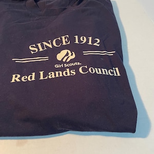 K&ouml;nnte beinhalten: Ein dunkelblaues T-Shirt mit wei&szlig;em Siebdrucktext, der SINCE 1912, das Girl Scouts-Logo und die Worte Red Lands Council zeigt. Das T-Shirt hat einen Rundhalsausschnitt und ein wei&szlig;es Etikett, das am Kragenbereich an der Innenseite sichtbar ist.