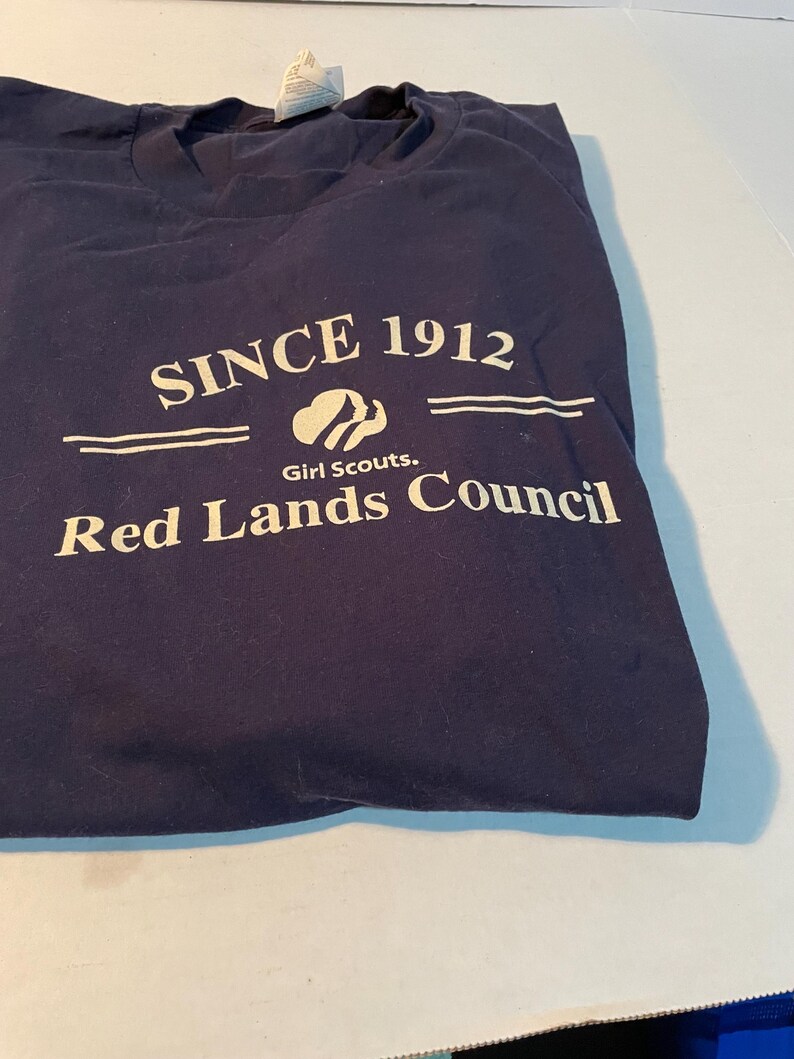 K&ouml;nnte beinhalten: Ein dunkelblaues T-Shirt mit wei&szlig;em Siebdrucktext, der SINCE 1912, das Girl Scouts-Logo und die Worte Red Lands Council zeigt. Das T-Shirt hat einen Rundhalsausschnitt und ein wei&szlig;es Etikett, das am Kragenbereich an der Innenseite sichtbar ist.