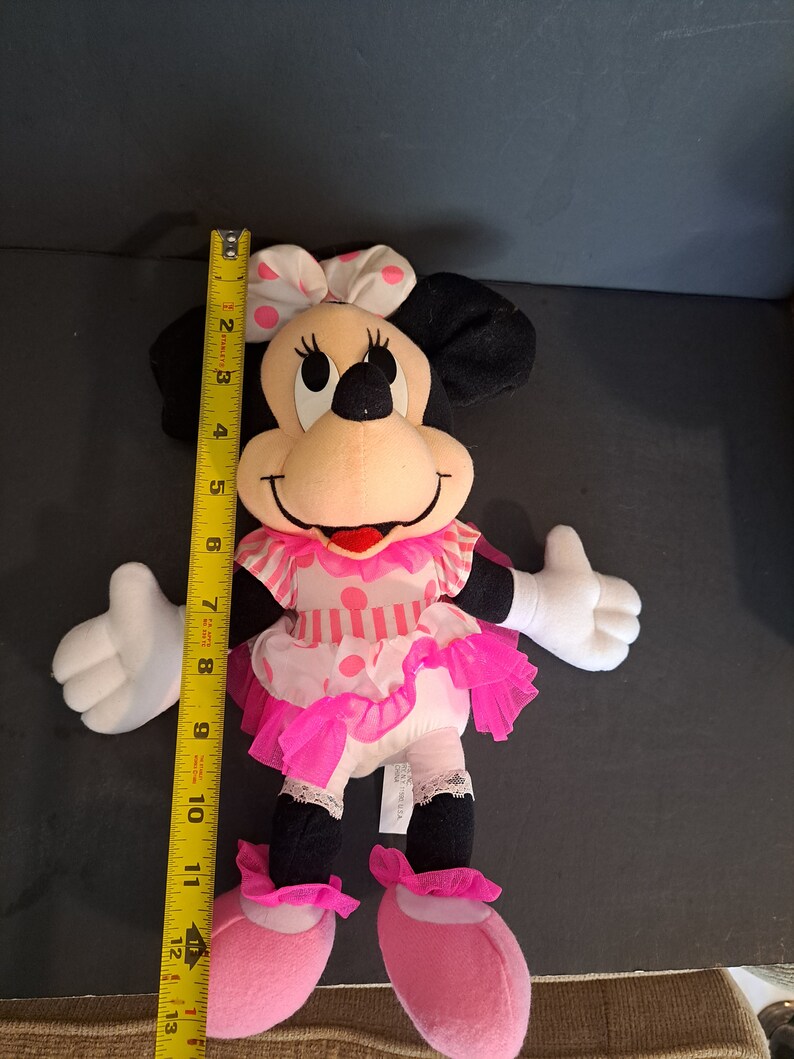 May include: Pink and black plush Minnie Mouse doll wearing a pink and white polka dot dress with a pink bow. The doll is approximately 12 inches tall.