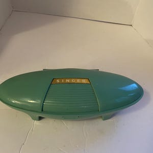 May include: Vintage teal Singer sewing machine case. The case is oval-shaped with a gold-colored panel that reads "SINGER". The case has a ribbed design on the front and is supported by four small feet.