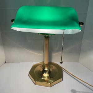 May include: A green-shaded desk lamp with a brass base and stem. The lamp has a pull chain and a white interior. The shade is a vibrant green color, and the base is octagonal.