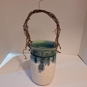 May include: A white ceramic vase with a green glaze, featuring a rustic twig handle. The vase is perfect for displaying flowers or greenery.