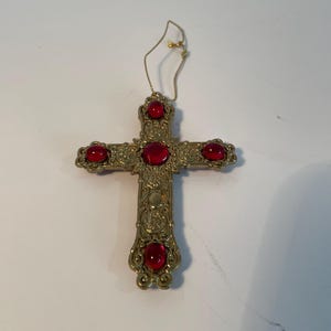 May include: Ornate gold-colored cross with five red jewel accents. The cross features intricate detailing and a loop for hanging. A decorative religious item.