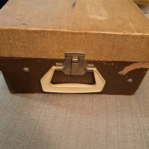 Pu&ograve; includere: Una valigia vintage marrone con una maniglia color crema e una chiusura in metallo. La valigia ha un esterno in tessuto testurizzato.