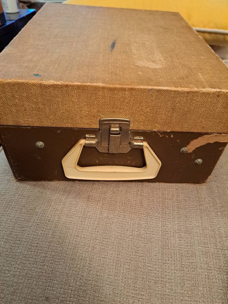 Pu&ograve; includere: Una valigia vintage marrone con una maniglia color crema e una chiusura in metallo. La valigia ha un esterno in tessuto testurizzato.