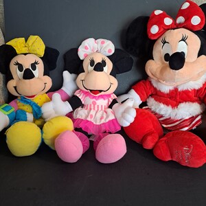 May include: Three plush Mickey Mouse characters. The character on the left is wearing a yellow and blue outfit with a yellow bow. The character in the middle is wearing a pink and white dress with a white bow. The character on the right is wearing a red and white outfit with a red bow.