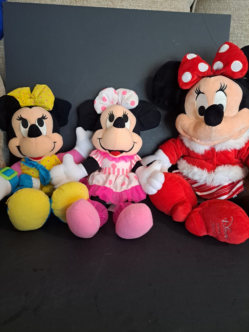 May include: Three plush Mickey Mouse characters. The character on the left is wearing a yellow and blue outfit with a yellow bow. The character in the middle is wearing a pink and white dress with a white bow. The character on the right is wearing a red and white outfit with a red bow.