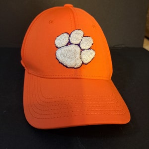 May include: An orange baseball cap with a white embroidered paw print. The paw print has a purple outline.
