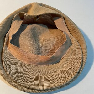 May include: A tan-colored flat cap featuring a woven, textured exterior and a light-colored interior sweatband. The cap has a rounded crown and a short, stiff brim, showcasing the detailed knit construction of the fabric from an interior perspective.