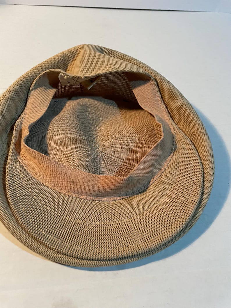 May include: A tan-colored flat cap featuring a woven, textured exterior and a light-colored interior sweatband. The cap has a rounded crown and a short, stiff brim, showcasing the detailed knit construction of the fabric from an interior perspective.