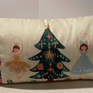 May include: A rectangular beige throw pillow featuring a printed illustration of a central dark green Christmas tree flanked by two nutcracker figures and two ballet dancers in yellow and blue dresses, all set against a textured fabric background.