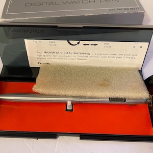 May include: A vintage Micronta Digital Watch/Pen in its original black case with an orange interior. The pen is silver-toned and rests on a beige foam insert. The case lid has text that reads "DIGITAL WATCH PEN".